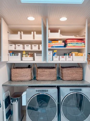 Laundry room with storage cabinets, towels, detergents, and baskets above a washing machine and dryer.