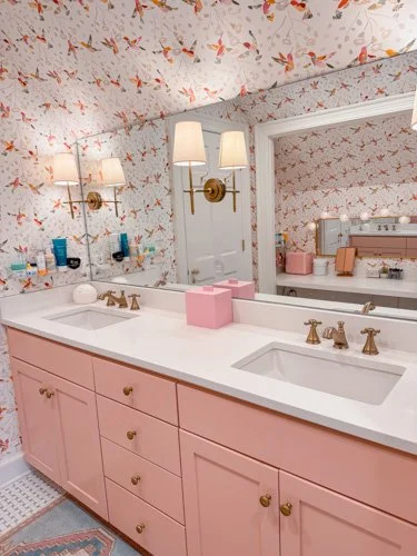 A bathroom with a pink vanity, double sinks, gold fixtures, a large mirror, wall-mounted lights, and floral wallpaper.