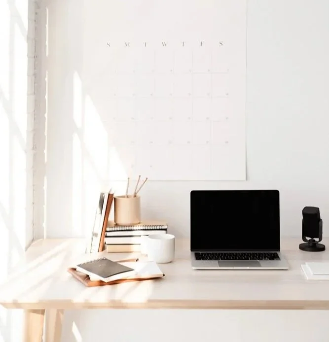 A clean, minimalist workspace with a wooden desk, an open laptop, a black speaker, a small stack of books, notebooks, a white mug, a beige container with pens, and a calendar on the wall in natural sunlight.
