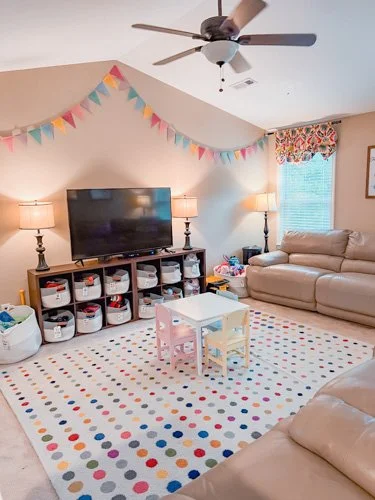 Living room with beige couches, a colorful polka dot rug, a large TV on a stand, two lamps, a small pink and yellow table, a window with a floral valance, and a multicolored pennant banner hanging from the ceiling.