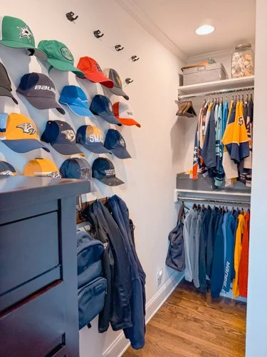 A closet with sports jerseys hung on racks, multiple baseball caps arranged on a wall, and backpacks on the floor.