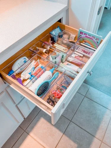 Open bathroom drawer filled with personal care items including lotions, hairbrushes, makeup, and toiletries.