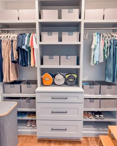 A neatly organized walk-in closet with clothing hanging on both sides, storage boxes on shelves, and three helmets on a middle shelf.