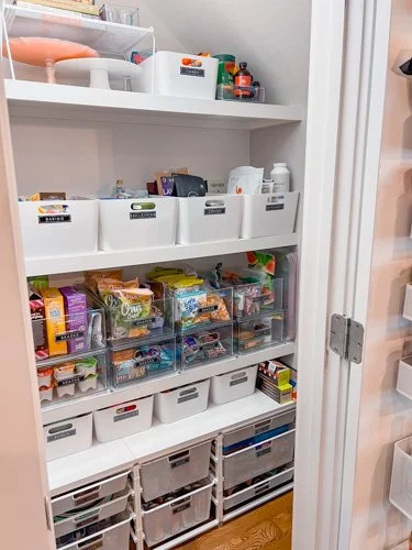Organized pantry with white shelves and clear plastic bins filled with snacks and food items.