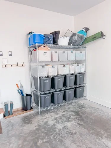 A metal shelving unit with three shelves filled with plastic storage bins in a garage or storage room. The top shelf holds various items, including a blue container and a green yoga mat. The middle and bottom shelves contain clear and gray bins. Lean