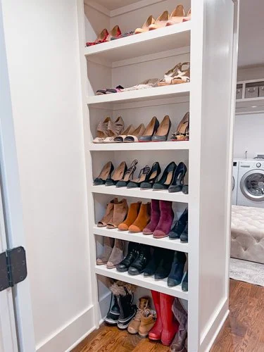 White bookshelf filled with various pairs of shoes, including heels, boots, and sneakers, in a laundry room or closet.