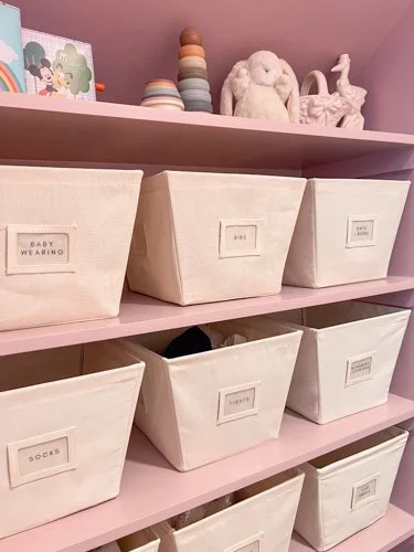 Pink shelf with white fabric storage bins labeled for baby items such as baby clothing, socks, and shoes. Plush toys and decorative items are on the top shelf.