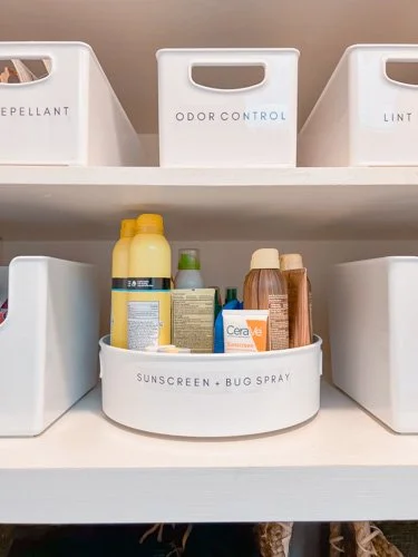 Bathroom shelf with storage bins labeled 'Repellent,' 'Odor Control,' and 'Linen.' Small bottles and spray containers in a round basket labeled 'Sunscreen + Bug Spray'.