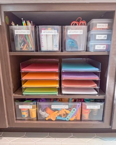 Wooden shelf with clear plastic storage bins containing art supplies, paper folders in various colors, and other miscellaneous craft items.