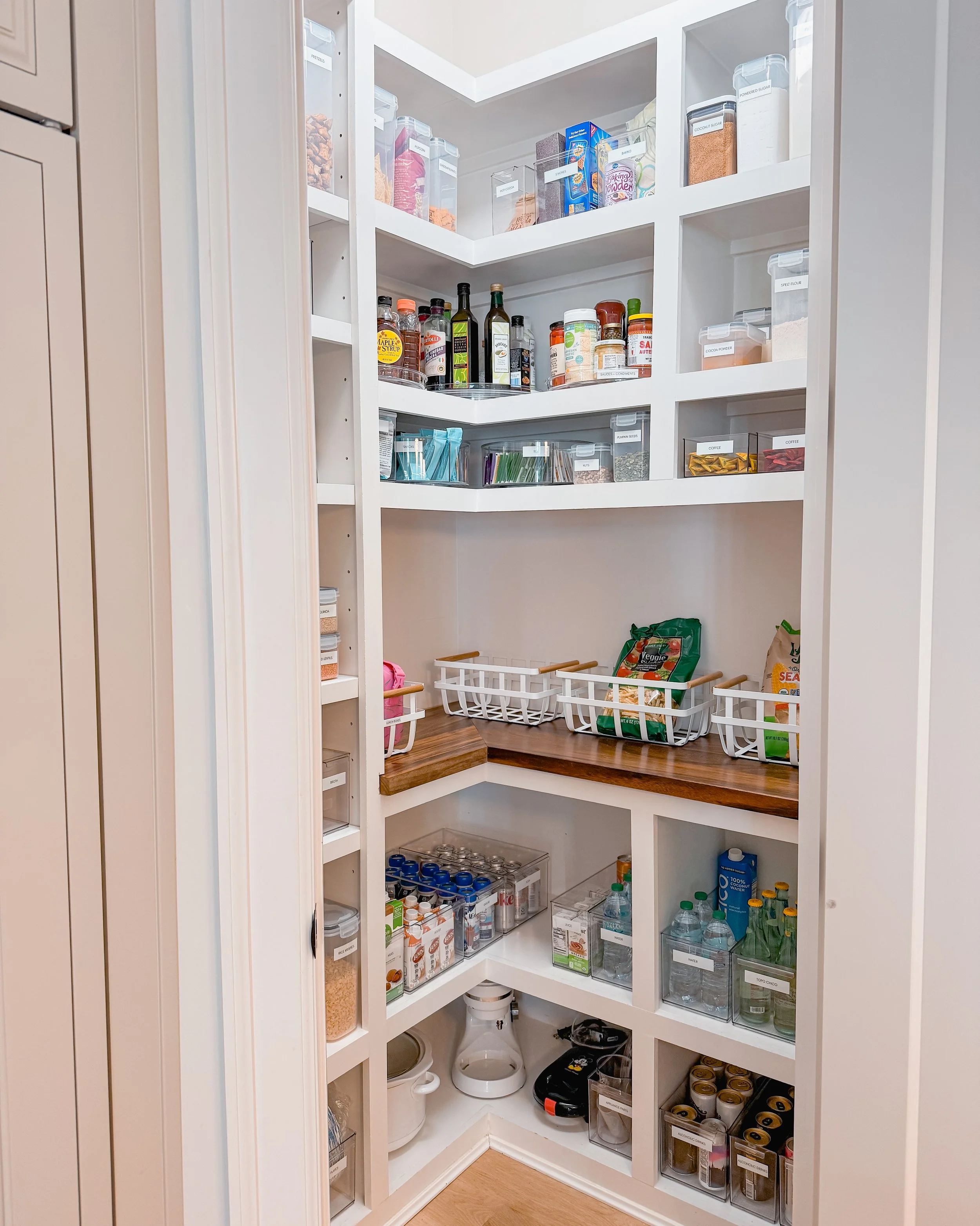 A well-organized pantry with white shelves stocked with various food and beverage items, including jars, bottles, boxes, and cans, with some empty baskets for storage.