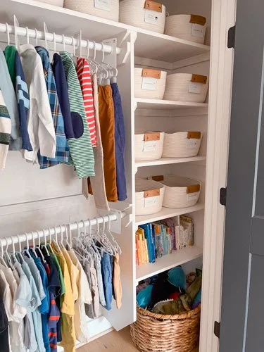Clothing closet with hanging shirts and pants, white storage baskets on shelves, and a wicker basket at the bottom containing shoes.