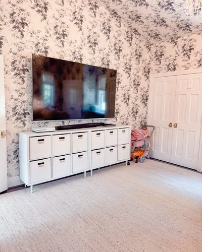 Living room with a large flat-screen TV on a white storage unit, floral wallpaper, white double closet doors, and a pink blanket on a cart.