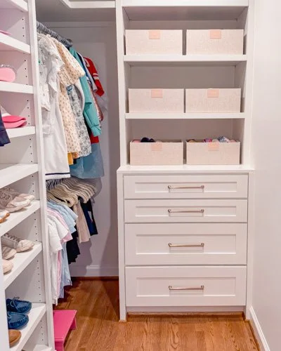 A clean walk-in closet with hanging clothes, shelves, and drawers, including storage bins on the top shelves.