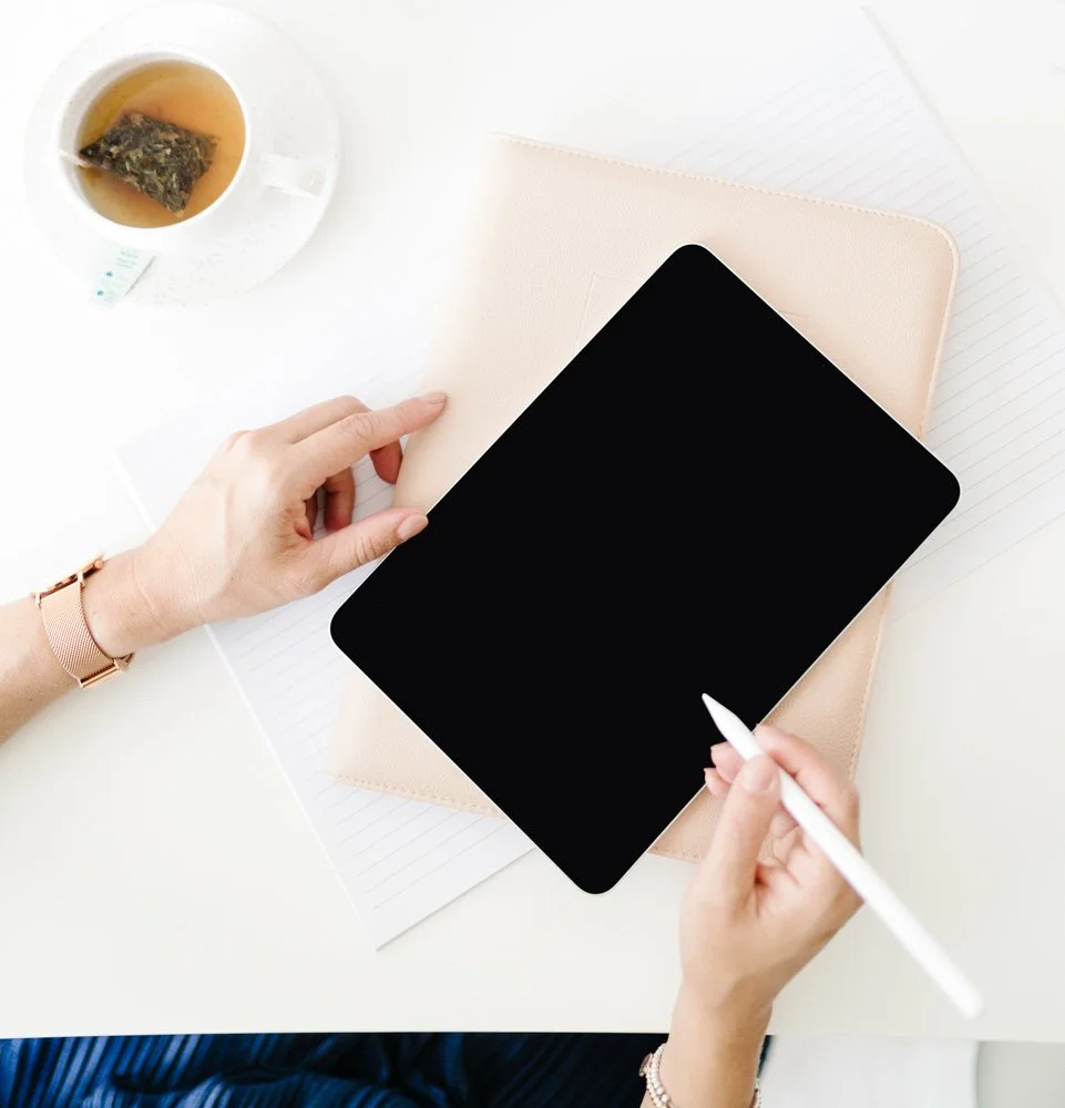 A person working with a black tablet, a beige notebook, and lined paper on a white desk. There is a cup of tea with a tea bag on a saucer nearby.