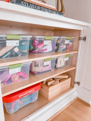 Shelves with multiple labeled plastic storage bins containing office and craft supplies, a wooden box, and a stack of books on top.