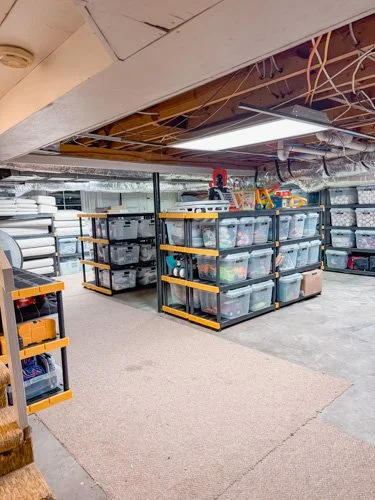 Storage area with plastic bins on black metal shelving units, some with yellow trim, in a basement with exposed ceiling pipes and wires.