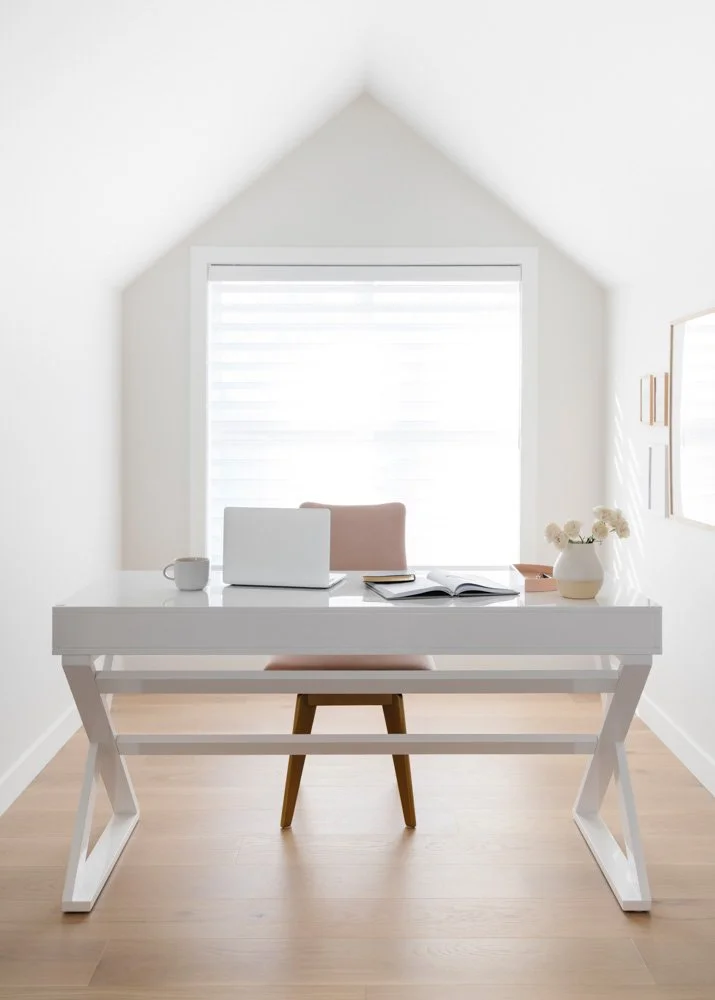 Bright, minimalist home office with white desk, pink chair, laptop, cup, open notebook, and vase of white flowers, with a window and simple wall decor.
