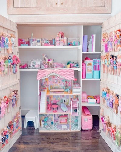 A dollhouse inside a closet with shelves, surrounded by numerous dolls on the closet walls, and a small stool.