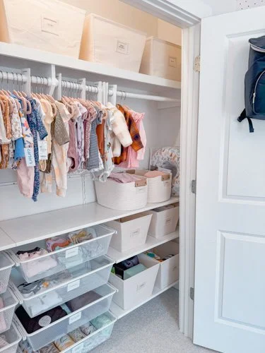 Organized closet with clothes on a hanging rail, storage bins and boxes on shelves, and a backpack hanging on the door.