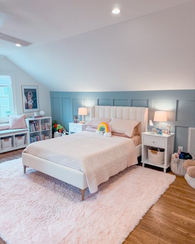 Bright bedroom with a white bed, pink and beige pillows, rainbow decoration, two white nightstands with lamps, bookshelf, and window with white blinds.