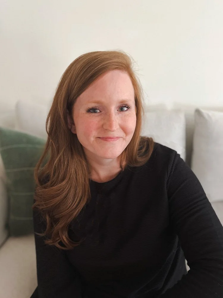 A woman with long red hair and a black top smiles while sitting on a light-colored couch with green and white pillows.