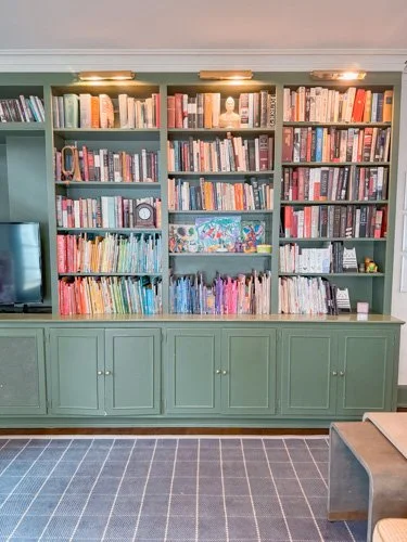 Green built-in bookshelf filled with books and decor, with a cabinet below, in a room with a carpeted floor and a small part of a chair visible in the corner.
