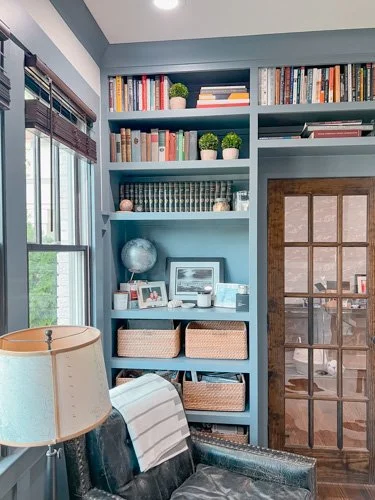 A blue built-in bookshelf filled with books, decorative plants, and framed photos, next to a window with blinds and a sliding glass door, in a cozy living room.