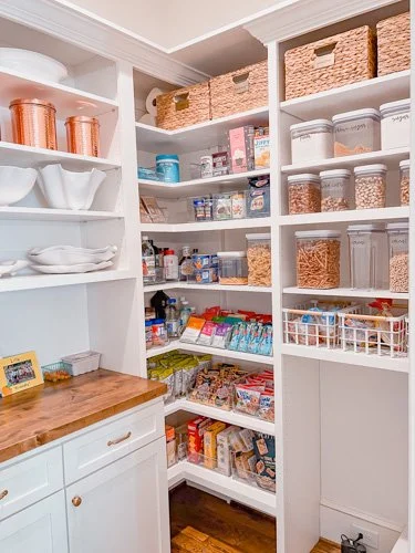 A well-organized pantry with white shelves filled with glass jars, baskets, canned goods, snacks, and kitchen items.