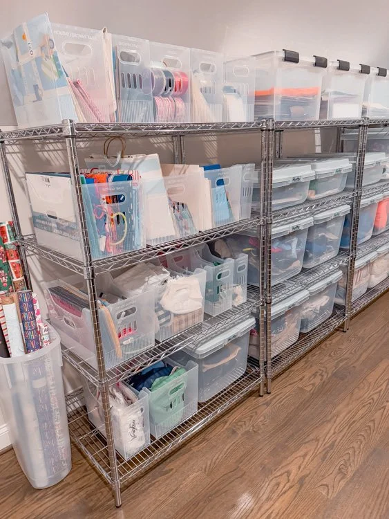 Clear storage bins and plastic baskets organized on metal shelves, holding various supplies, against a light-colored wall on hardwood floor.