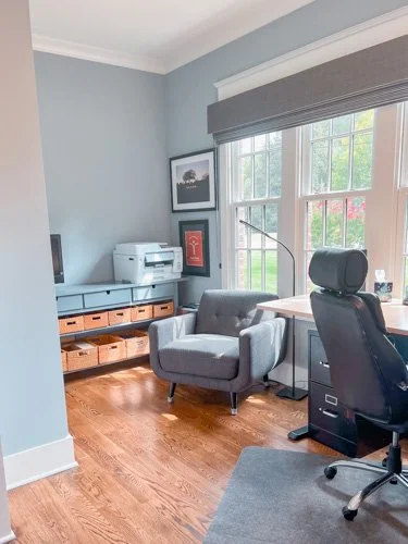 Home office with a gray armchair, a black desk chair, a printer on a wooden stand, a framed picture, and a large window with blinds letting in natural light.
