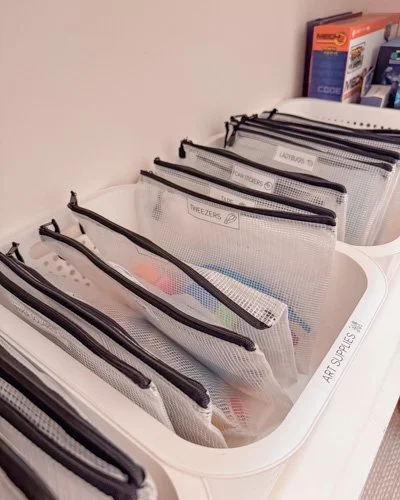 Clear mesh pouches organized in a white bin, labeled for storage of items like deodorants, freezer bags, and art supplies.
