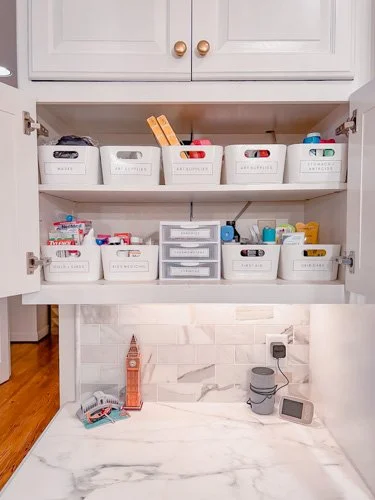 Open kitchen cabinet with white storage bins containing various small items, a small plastic drawer unit, and a marble countertop featuring a miniature Big Ben clock, a roll of paper towels, and a digital timer.