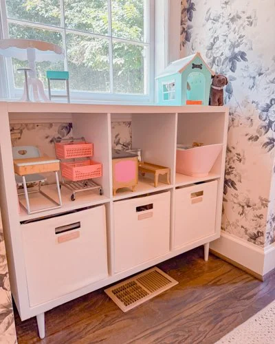 A white storage cabinet with three sections and six drawers, decorated with a dollhouse, a pink plastic basket, and small cushions, situated against a floral wallpapered wall near a window.