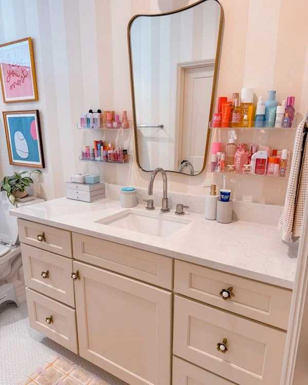 A tidy bathroom vanity with a large mirror, organized skincare products on shelves, and pastel decor on the wall.