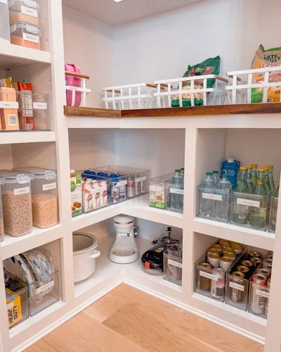 Pantry with open white shelves featuring jars of grains, snacks on a wooden counter, and small kitchen appliances in a corner.