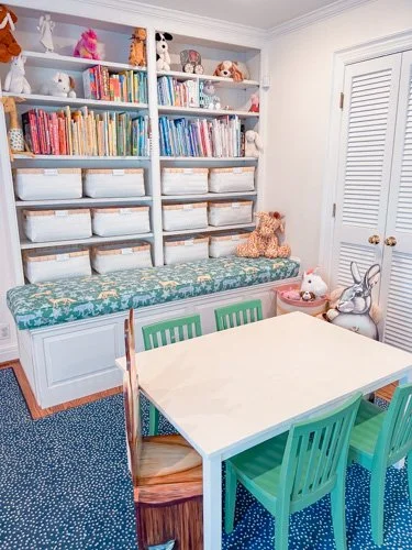 A cozy kid's room with a white bookshelf filled with books, stuffed animals, and storage bins, a cushioned bench with a floral cushion, a white table surrounded by four green chairs, and toys including a rabbit-shaped plush on the floor.