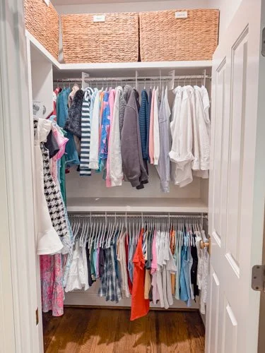 Inside a closet with hanging clothes and storage baskets on top.