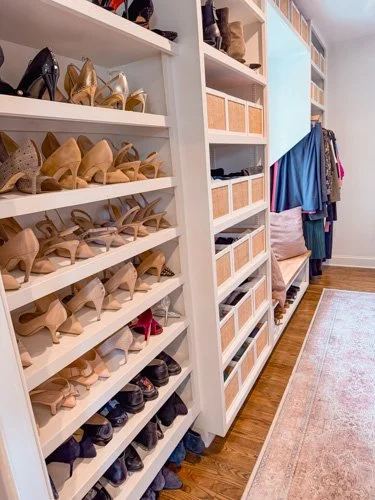 Walk-in closet with white shelves filled with shoes and baskets, a clothing rack with hanging clothes, and a beige area rug on hardwood floor.