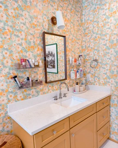 Bathroom vanity with a white countertop, brown cabinets, a large mirror, and shelves with toiletries, with floral wallpaper.