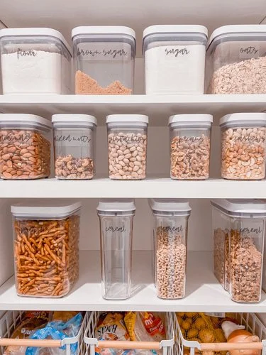 White shelves with clear labeled containers filled with flour, brown sugar, sugar, oats, nuts, and snacks. Additional plastic containers on lower shelves hold various snacks and supplies.