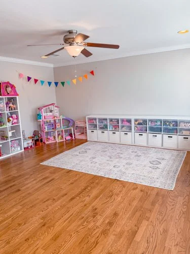 Children's playroom with colorful storage shelves and a rainbow banner on the wall, a ceiling fan, and a light-colored area rug on hardwood floors.