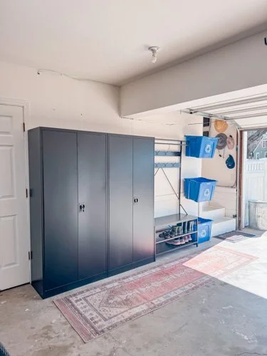 Garage with a black storage cabinet, blue bins, and sports equipment near an open garage door.