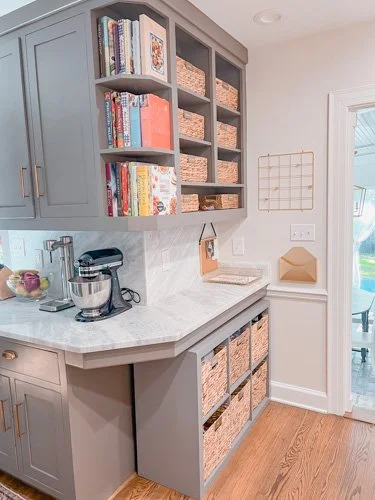 Kitchen corner with gray cabinets, open shelves, brown baskets, cookbooks, and kitchen appliances on a white marble countertop.