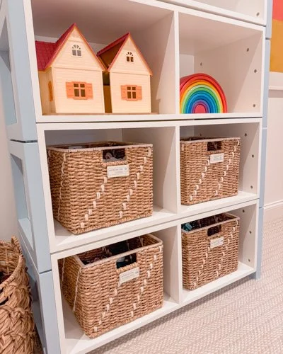 A white shelving unit with three rows of storage compartments. The top row contains two small dollhouses and a rainbow-shaped object. The middle and bottom rows have woven storage baskets labeled with small white tags.