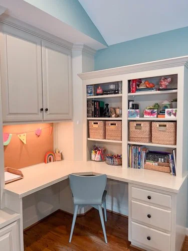 Home office corner with built-in white desk, a light blue chair, and a white bookshelf filled with books and storage baskets, with light blue walls.