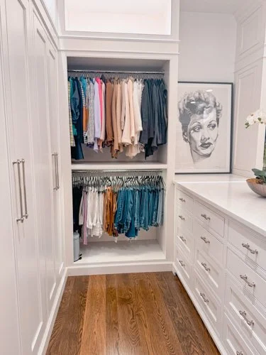 Walk-in closet with white built-in shelves filled with hanging clothes in various colors, a framed black-and-white portrait, and a white cabinet with drawers on the right side.