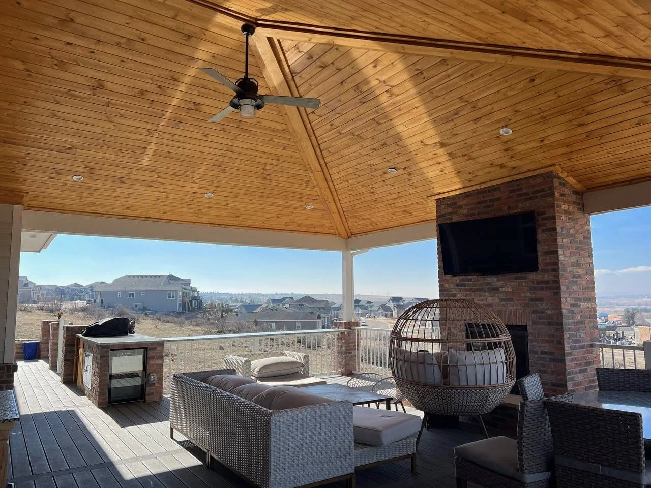 Covered outdoor patio with a wooden ceiling, ceiling fan, brick fireplace with a mounted TV, wicker chair with cushions, outdoor furniture, and a view of neighboring neighborhood houses.