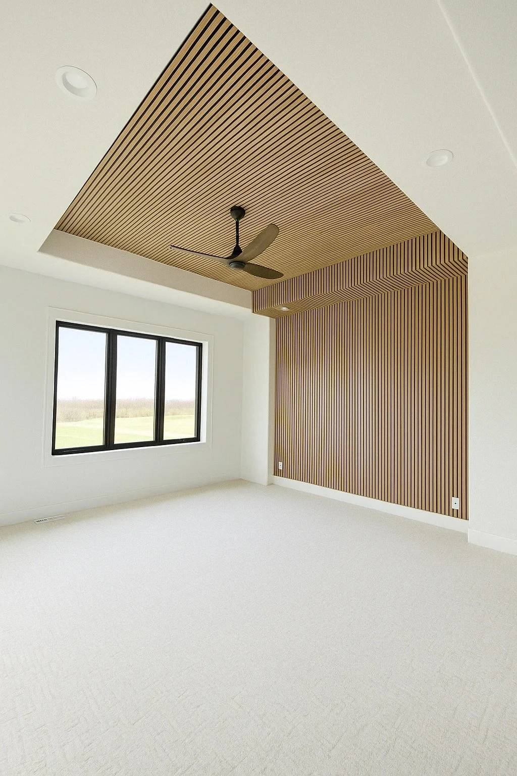Empty room with white walls, beige carpet, a large window showing an outdoor view, and a ceiling with wooden panels, a black fan, and recessed lighting.