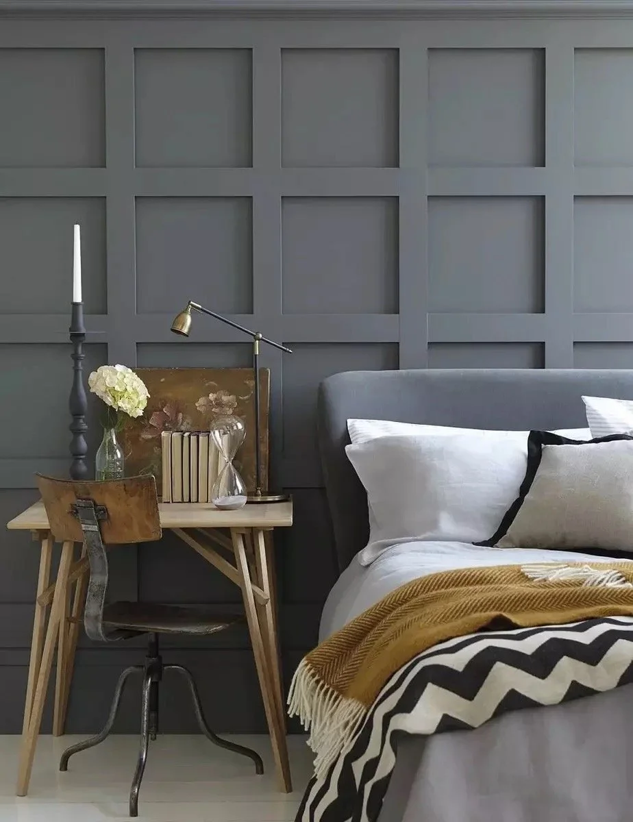 A bedroom corner featuring a bed with white and beige pillows, a mustard yellow blanket, and a black and white zigzag throw. Next to the bed is a small, light wood table with a glass vase holding white flowers, a stack of books, an hourglass, and a b