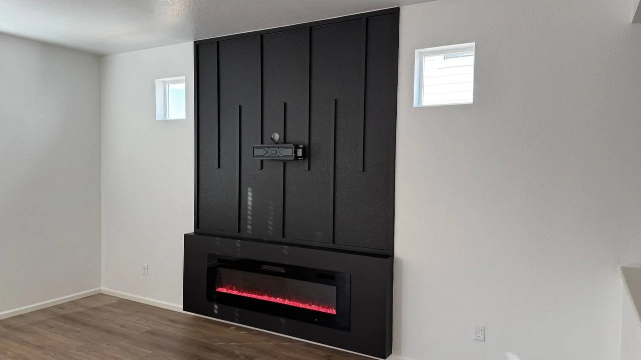 Modern interior with a white wall, two small windows, and a black wall-mounted electric fireplace with red flames, above which is a black panel with a TV mount.